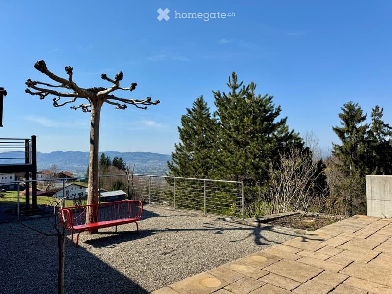 Outdoor patio with a red bench, gravel surface, metal railing, and scenic mountain view.