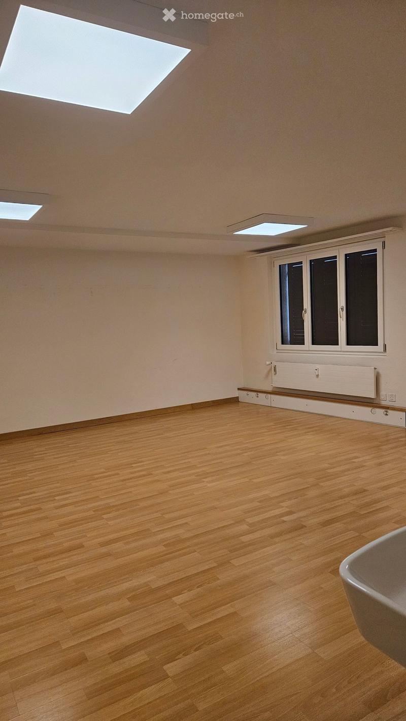 Empty room with wooden flooring, white walls, and large windows with blinds. The room has several ceiling lights and a radiator under the window.