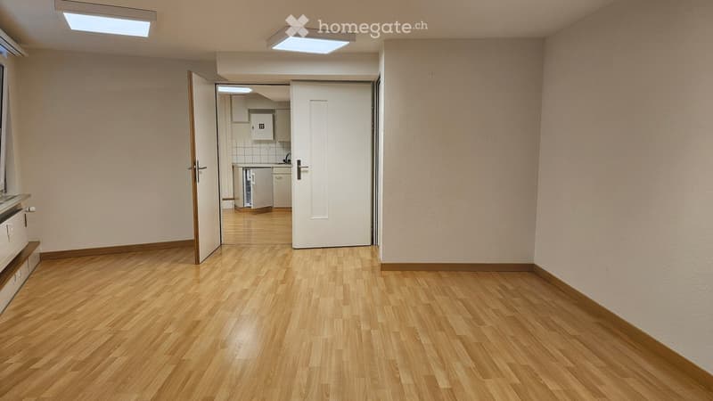 Empty room with wooden flooring, light-colored walls, and a door leading to a small kitchen area.