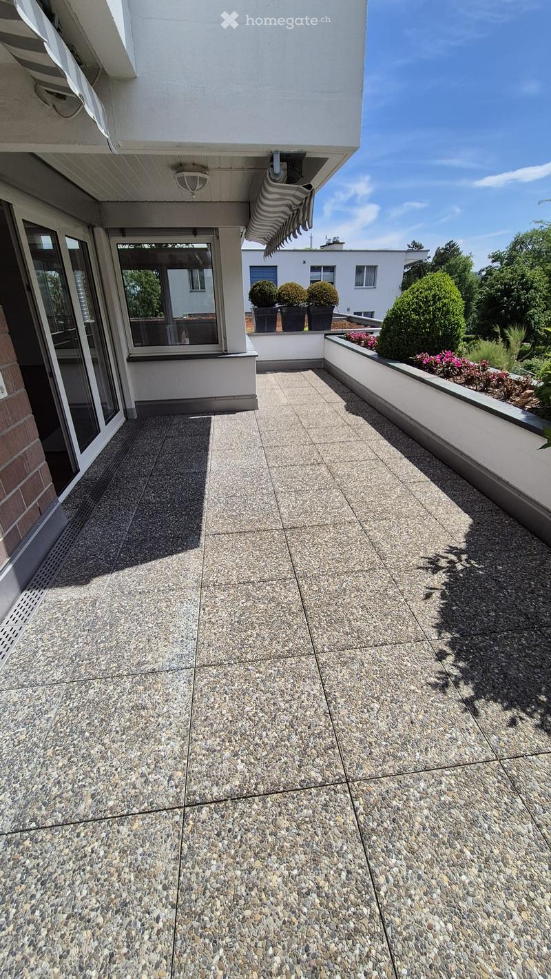 Balcony with tiled floor, glass door leading to interior, small garden area with potted plants and flowers, and a striped towel hanging from the building.