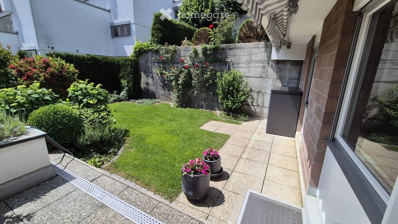 Small, well-maintained garden with a neatly trimmed lawn, surrounded by various shrubs and potted plants. The area is bordered by a low concrete wall and features a paved pathway leading to a covered patio area.