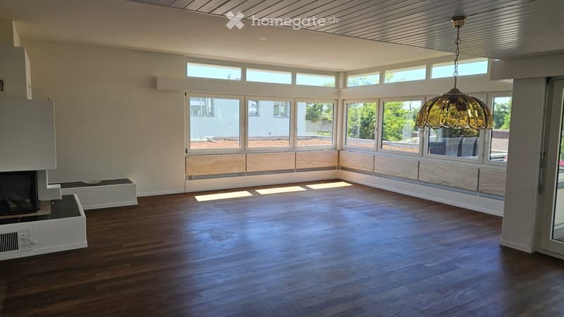 Bright, empty room with large windows, wooden flooring, and a modern fireplace. No furniture or furnishings are present.