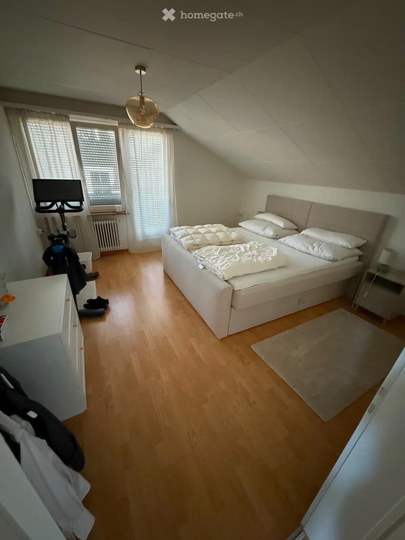 Bright bedroom with wooden flooring, large window with white curtains, bed with white bedding, nightstand with lamp, exercise bike, and mirror.