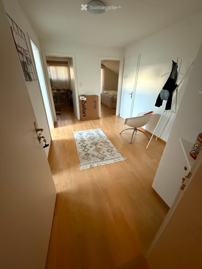 Bright hallway with wooden flooring, white walls, and a small rug. Visible doors lead to other rooms, and a coat rack with clothes is on the right side.