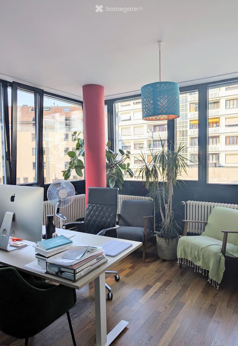 A modern, well-lit room with large windows, a desk with a laptop, books, and a fan, along with a comfortable seating area featuring a sofa and a plant.