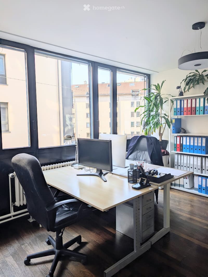 Modern office space with a large wooden desk, ergonomic chair, dual monitors, laptop, desk lamp, potted plant, and shelves with binders and office supplies.