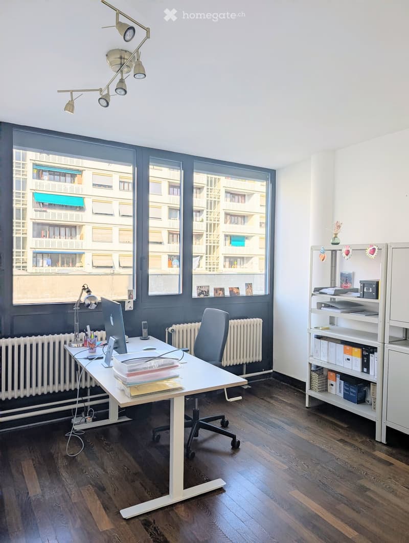 Modern office space with a white desk, ergonomic chair, laptop, bookshelf, and decorative vase. Large windows offer a city view.