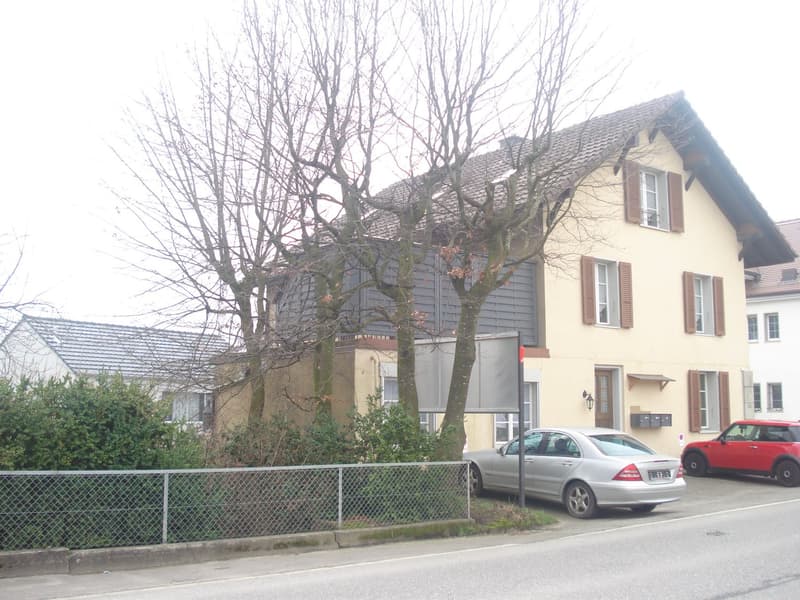 Two-story house with yellow facade, multiple windows, and a small balcony. There are two parked cars in front of the house, and a fence lines the front yard.