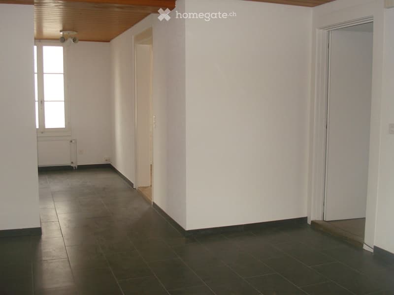 Empty hallway with tiled floor, white walls, and wooden ceiling. Features include a window with blinds, a radiator, and a door leading to another room.