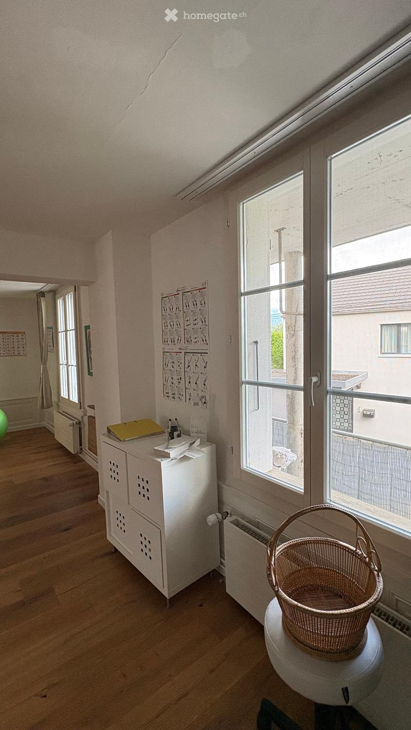 A small, well-lit room with wooden flooring, a white cabinet with a yellow folder, a green ball, and a wicker basket on a stool near a large window with a view of the building exterior.
