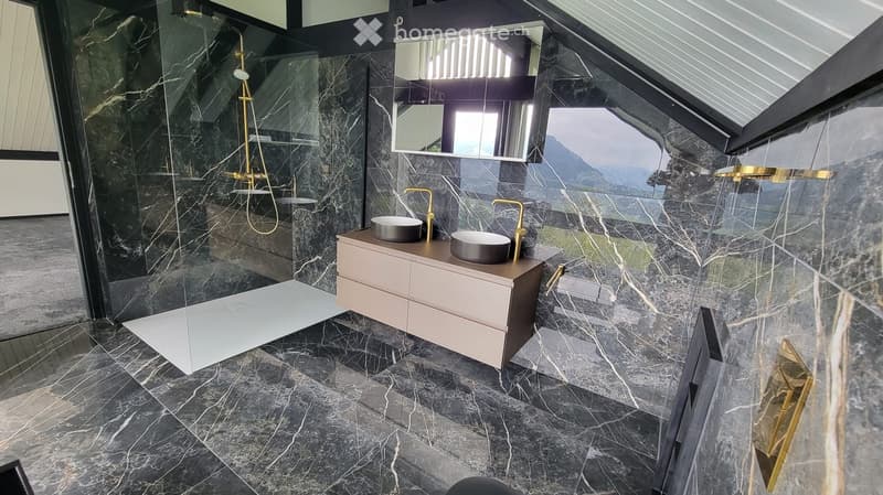 Modern bathroom with dark marble tiles, double sink vanity, gold fixtures, and a glass-enclosed shower area.