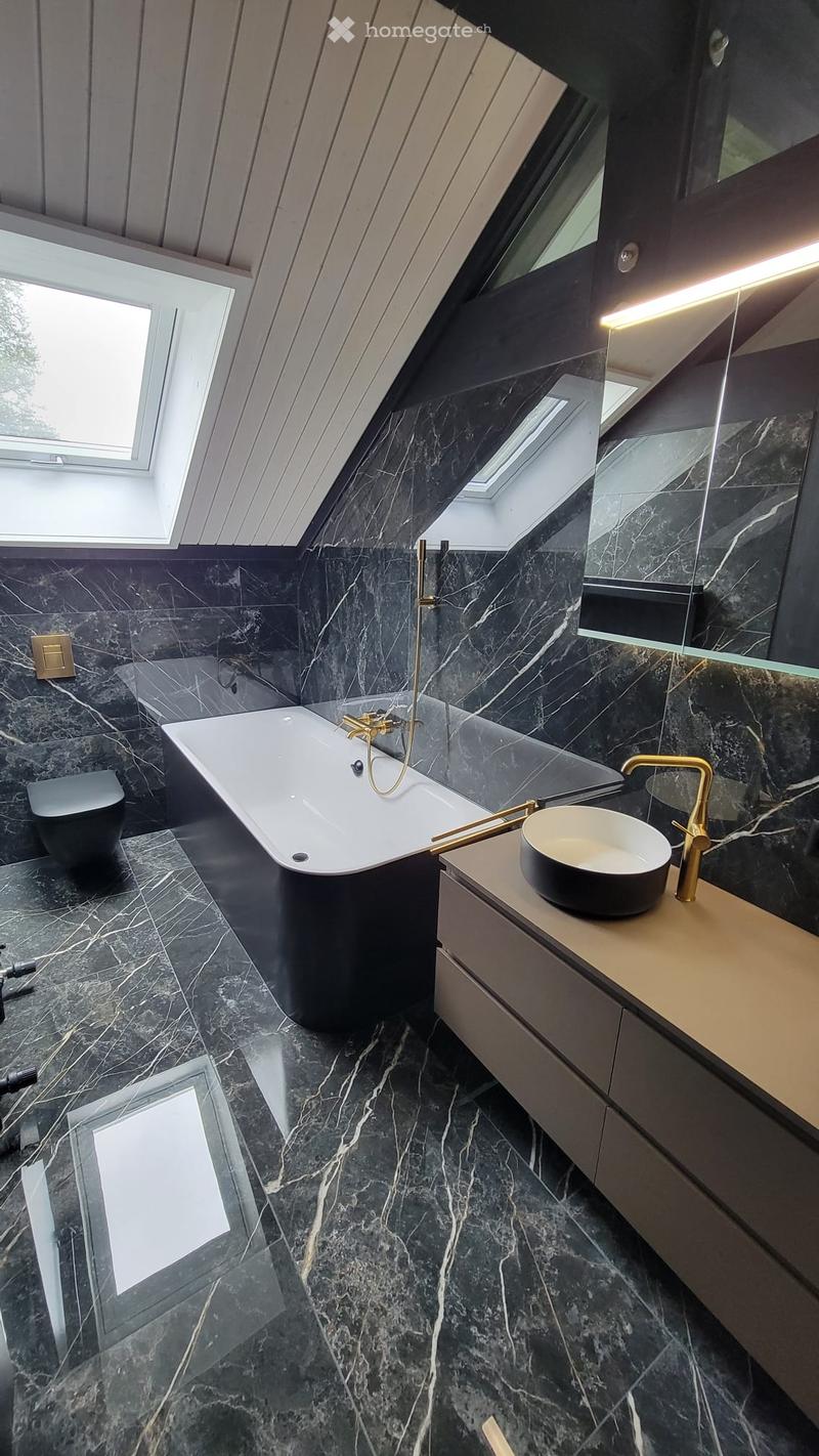 Modern bathroom with black marble tiles, a freestanding bathtub, a sink with a gold faucet, and a large mirror. The room features skylights and a sleek, contemporary design.