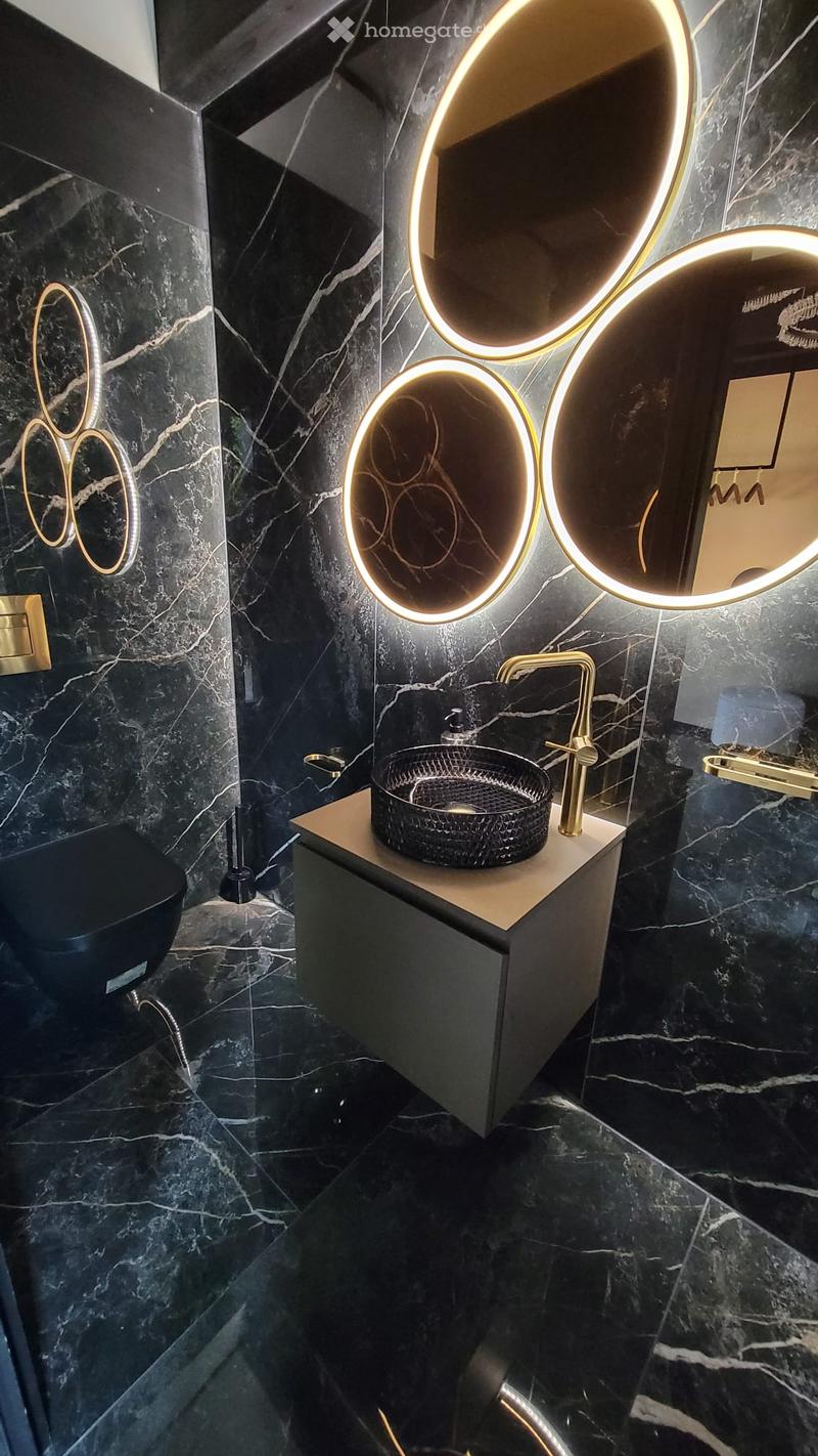 Modern bathroom with black marble walls and floor, a sleek sink with a gold faucet, a black woven sink basin, and a round illuminated mirror above the sink. A toilet is visible to the left.