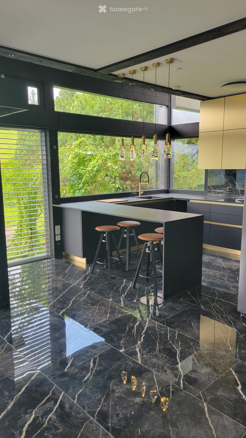Modern kitchen with a central island, four bar stools, sleek cabinets, and pendant lights. The floor is black marble with a striking veined pattern, and the space features large windows with a view of greenery.
