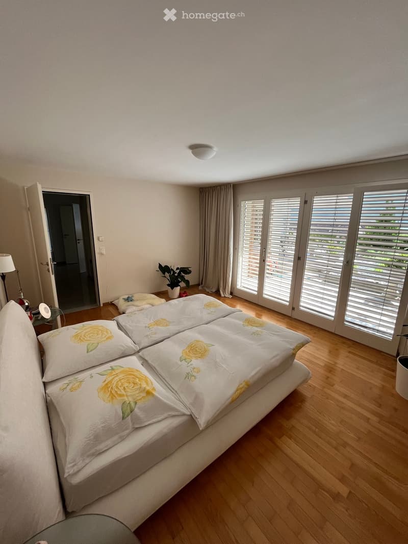 Bright bedroom with wooden flooring, large bed with yellow rose-patterned bedding, bedside table with lamp, plant, and large windows with blinds.