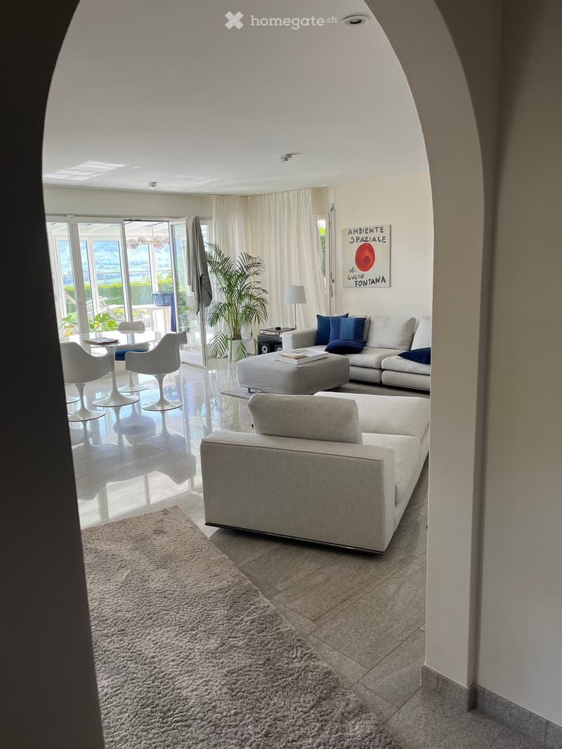 Modern living room with large windows, white sofa, ottoman, chairs, coffee table, lamp, and a plant near the window.