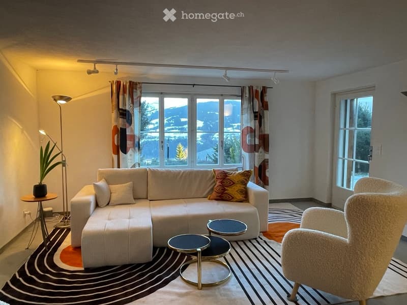 Modern living room with large windows offering a scenic view, featuring a white sectional sofa, decorative pillows, a round coffee table, a floor lamp, and a potted plant.