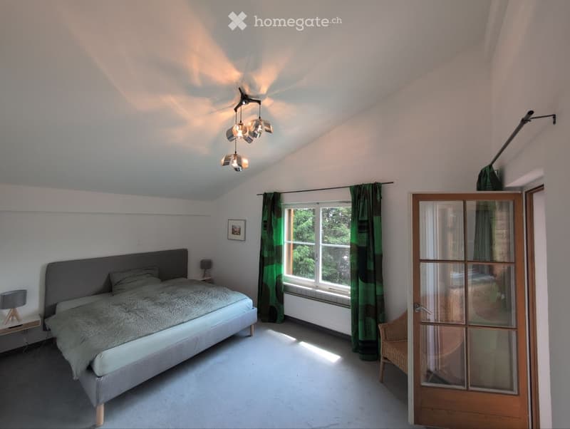 Bright bedroom with a grey bed, nightstand, lamp, and a large window with green curtains. A wicker chair is near the door.