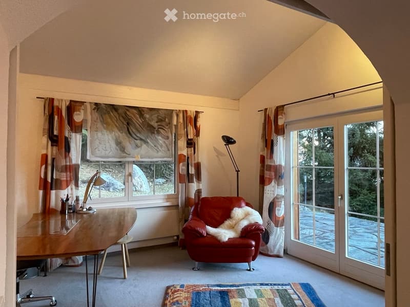 A cozy room with a slanted ceiling, large windows, and a sliding glass door leading to an outdoor area. Features include a wooden table, red leather chair with a fur throw, a floor lamp, and colorful curtains.