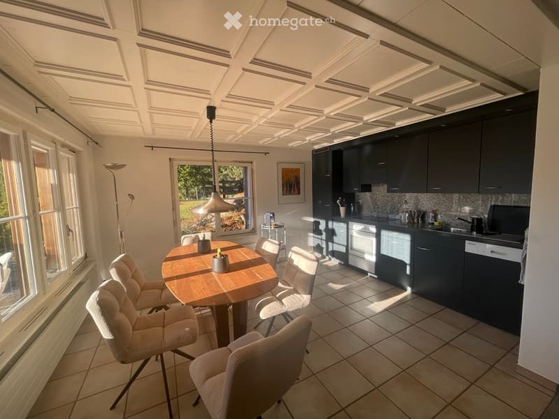 Bright, modern kitchen and dining area with dark cabinets, a wooden dining table, white chairs, and a hanging pendant light. The kitchen features stainless steel appliances, including an oven, dishwasher, and sink.