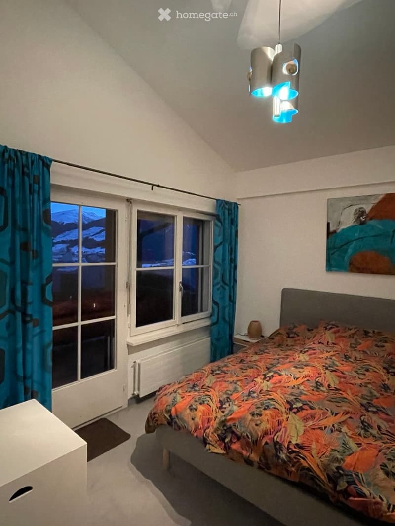 Cozy bedroom with a large bed, colorful bedspread, nightstand, and a painting on the wall. Large windows with blue curtains offer a view of the night sky.