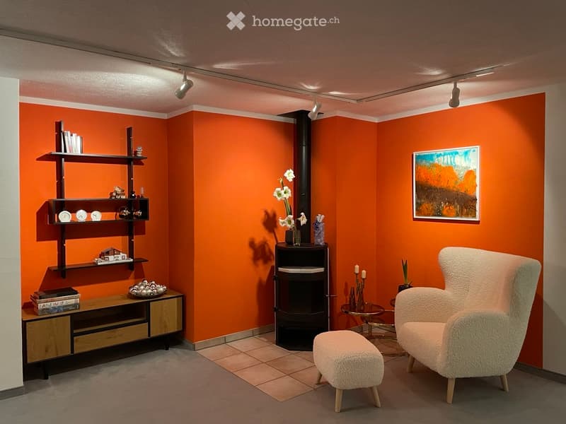 Modern living room with orange walls, a fireplace, a white armchair, a stool, a shelf with decorative items, and a small table with candles.