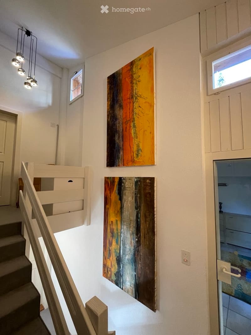 Staircase with modern wooden railing, colorful abstract paintings on walls, pendant lights, and a view into an adjacent room.