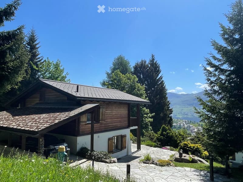 A two-story wooden house with a tiled roof, surrounded by trees and mountains, with a driveway and garden area.
