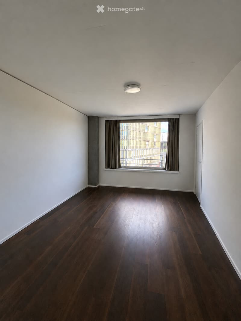 Empty room with wooden flooring, white walls, and a large window with curtains. The room has a single ceiling light fixture and a door on the right side.
