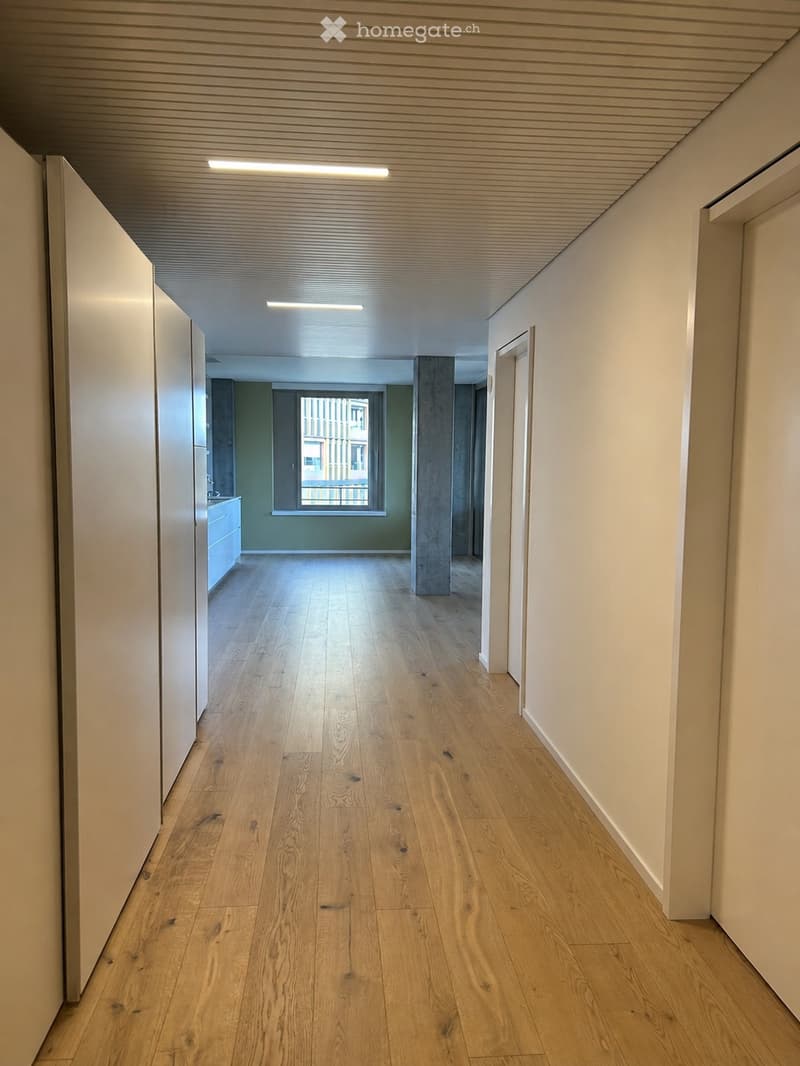 Empty hallway with light wooden flooring, white walls, and recessed lighting. A window at the end provides natural light, and a small room with a sink is visible to the left.