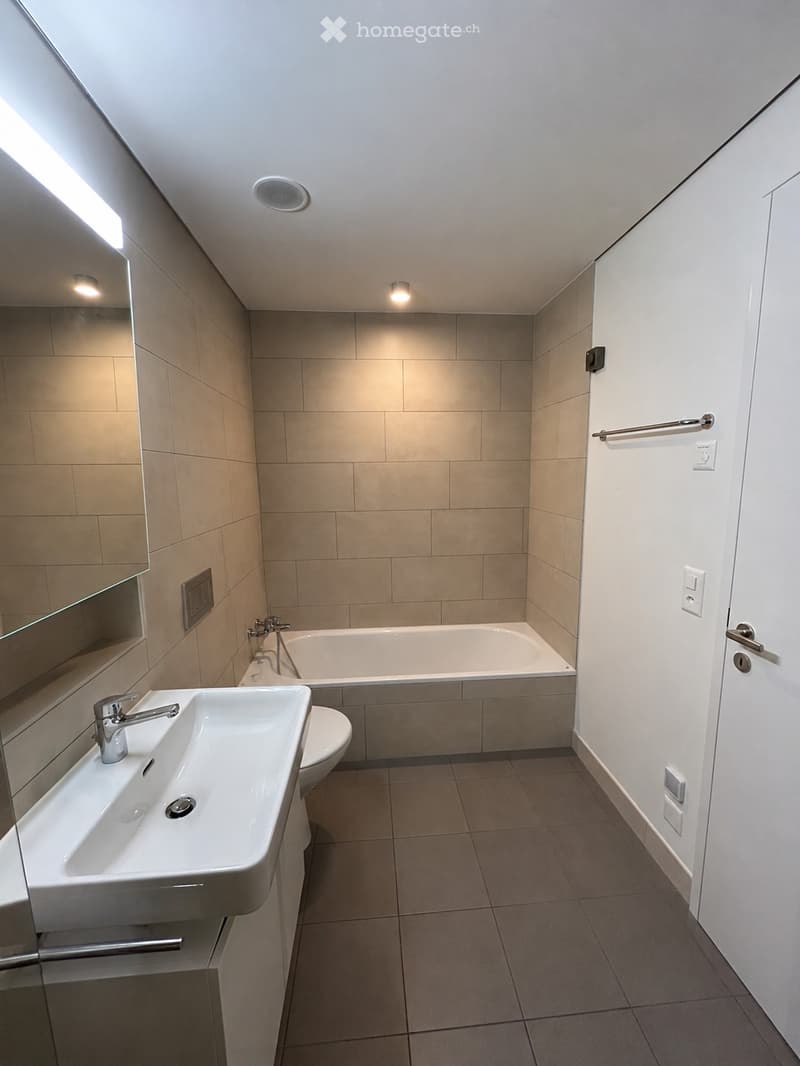Modern bathroom with beige tiled walls and floor, featuring a white sink with a chrome faucet, a bathtub, a toilet, and a large mirror with built-in storage. The room includes recessed lighting and a towel rack on the wall.