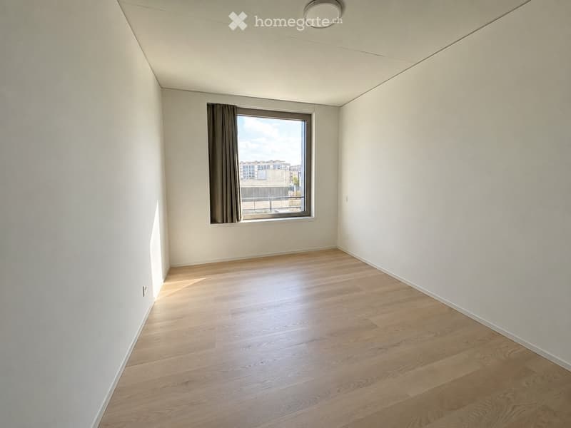 Empty room with light wooden flooring, white walls, and a large window with a curtain, offering a view of the city.