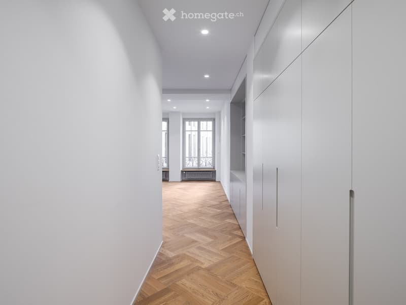 Modern hallway with herringbone wooden flooring, white walls, built-in storage units, and recessed lighting. The hallway leads to a room with large windows and a radiator.