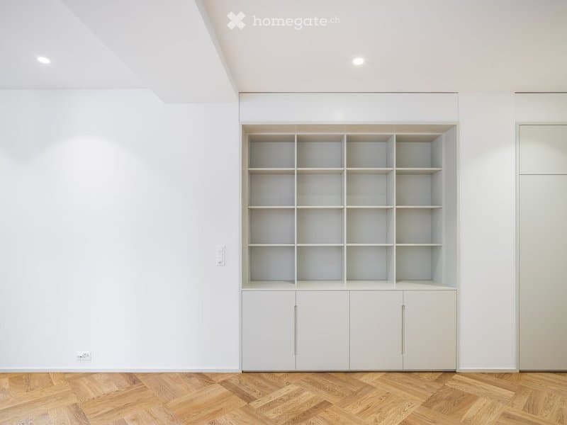 Empty room with white walls, wooden herringbone floor, and built-in white shelving unit with multiple compartments and closed cabinets.