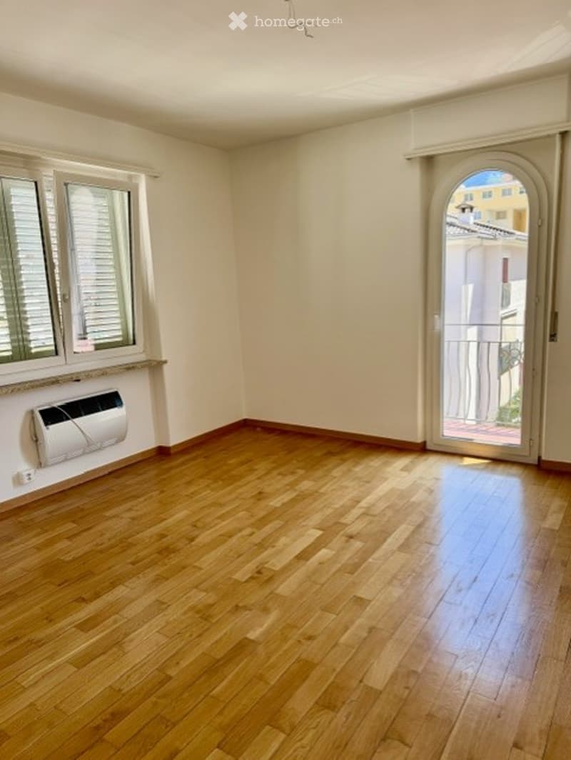 Empty room with wooden flooring, white walls, large windows with shutters, and a sliding glass door leading to a balcony.