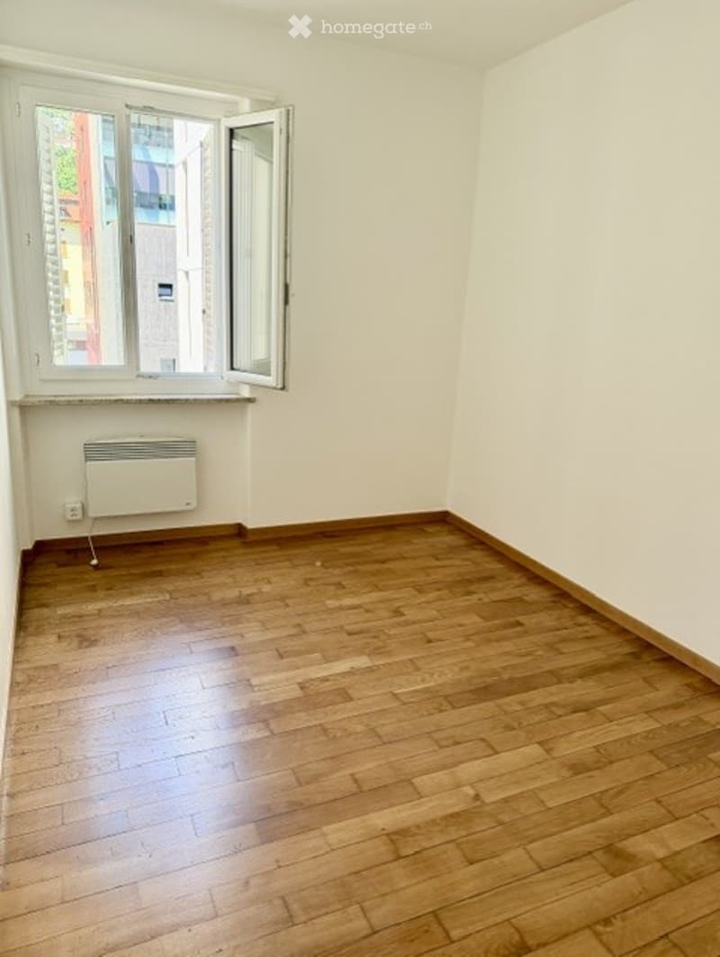 Empty room with wooden flooring, white walls, and a window with open shutters. A radiator is visible below the window.