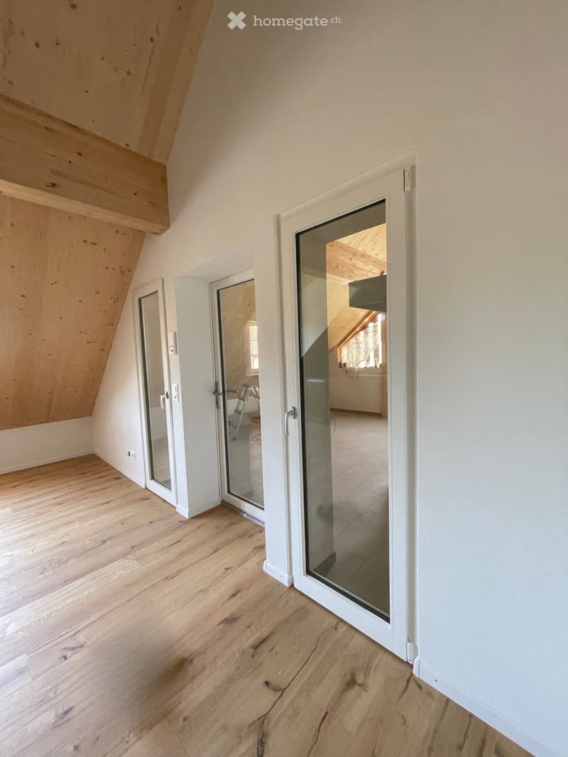 Empty attic room with wooden flooring, white walls, and large glass doors leading to another room.