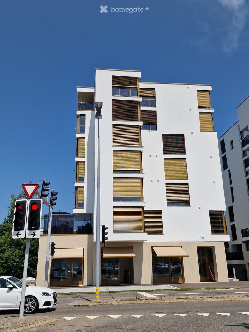 Modern multi-story building with white facade, large windows, and balconies. The ground floor features commercial spaces with glass doors and awnings. Traffic lights and a white car are visible in the foreground.