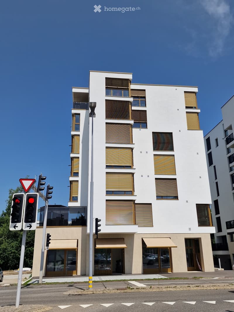 Modern multi-story building with white facade, large windows, and balconies. Features include a ground floor with commercial storefronts and traffic lights at the intersection.