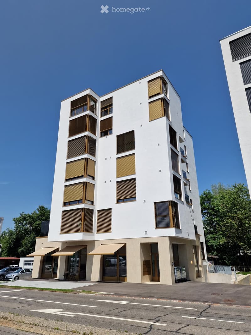 Modern multi-story building with white facade, large windows with wooden shutters, and a lower commercial section with awnings and glass doors.