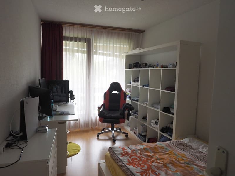 A well-lit bedroom with a bed, desk, gaming chair, and shelves filled with various items. The room features wooden flooring and large windows with curtains.