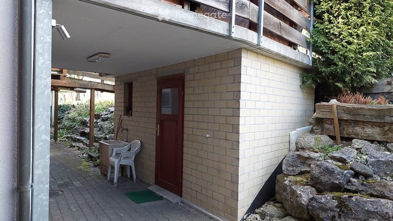 Small outdoor area with a tiled pathway, a red door, a white plastic chair, and a small garden with rocks and greenery.
