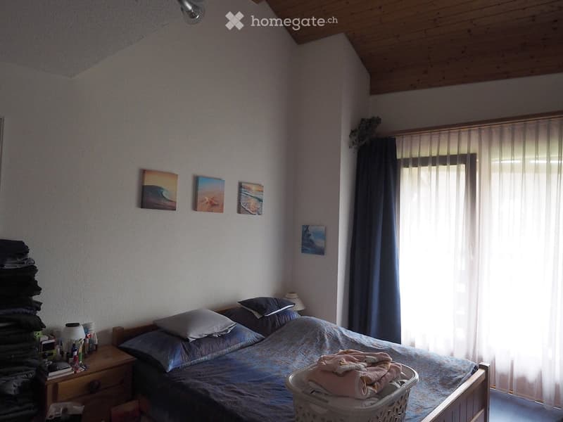Cozy bedroom with a double bed, nightstand, lamp, and artwork on the walls. A basket with folded blankets sits on the bed, and a stack of folded clothes is visible on the left side.