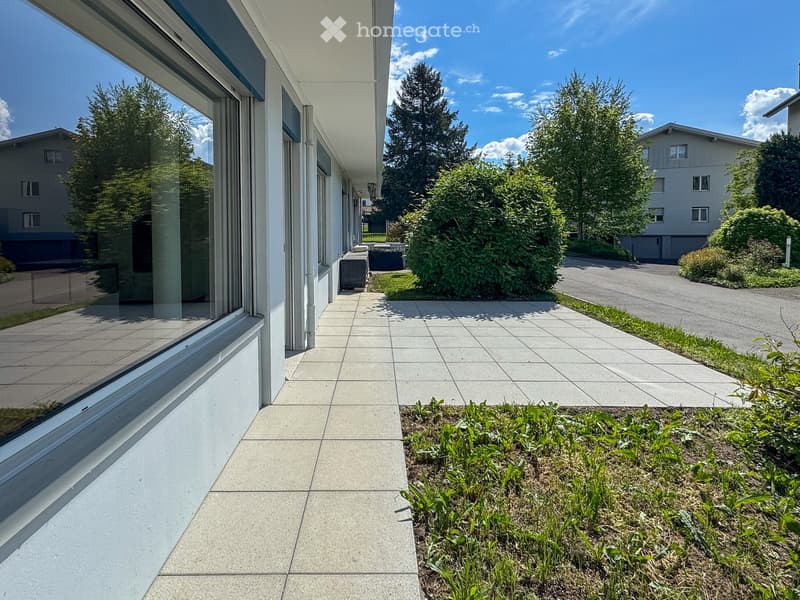 Balcony area with tiled flooring, modern building facade, greenery, and a paved driveway leading to neighboring houses.