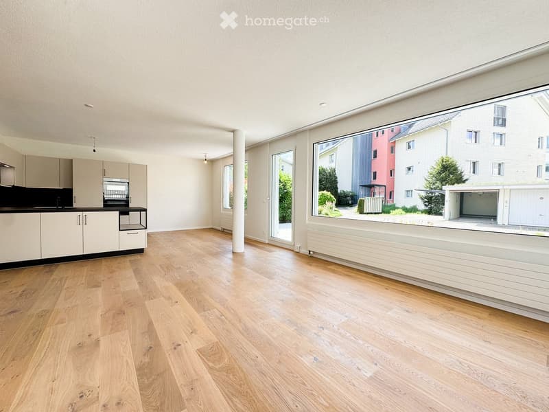 Open-plan unfurnished room with light wooden flooring, white walls, large windows, and a modern kitchen area with cabinets and a countertop.
