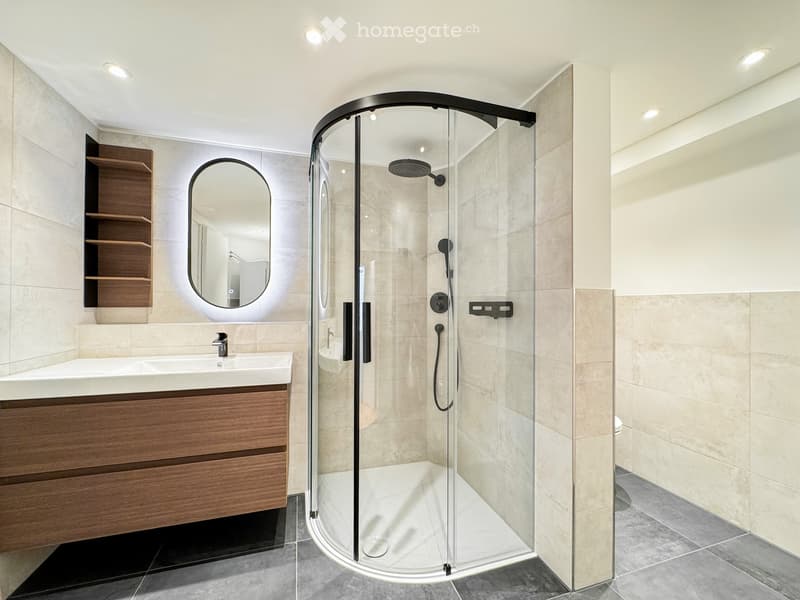 Modern bathroom with a glass-enclosed shower, a sink with a wooden cabinet, and a round mirror on the wall.