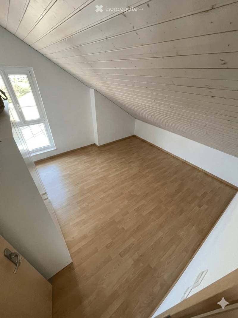 Empty attic room with wooden flooring, white walls, and a slanted wooden ceiling. A window on one side allows natural light to enter.