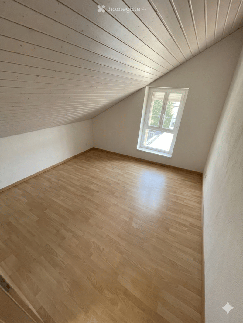 Empty attic room with wooden flooring, white walls, and a slanted wooden ceiling. A single window provides natural light.