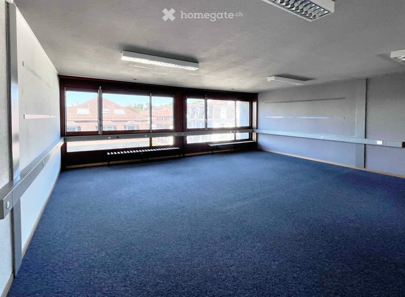 Empty room with blue carpet, large windows, and minimal furnishings including a small bench and a low cabinet.