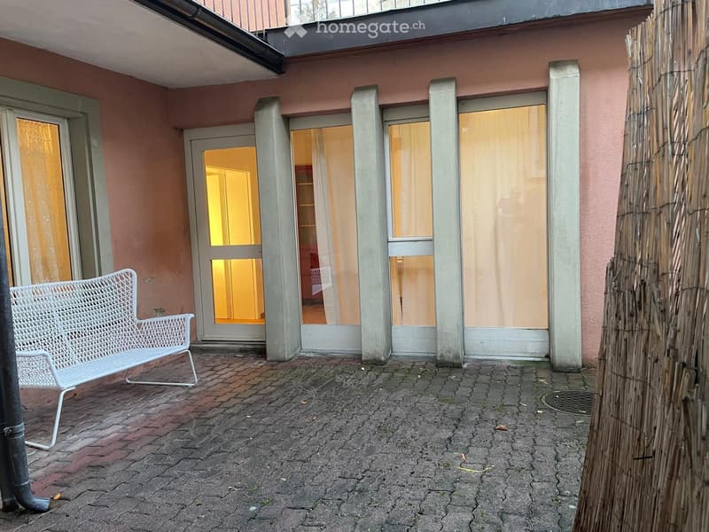 Balcony with brick floor, white metal bench, bamboo screen, and open doors leading to interior space.